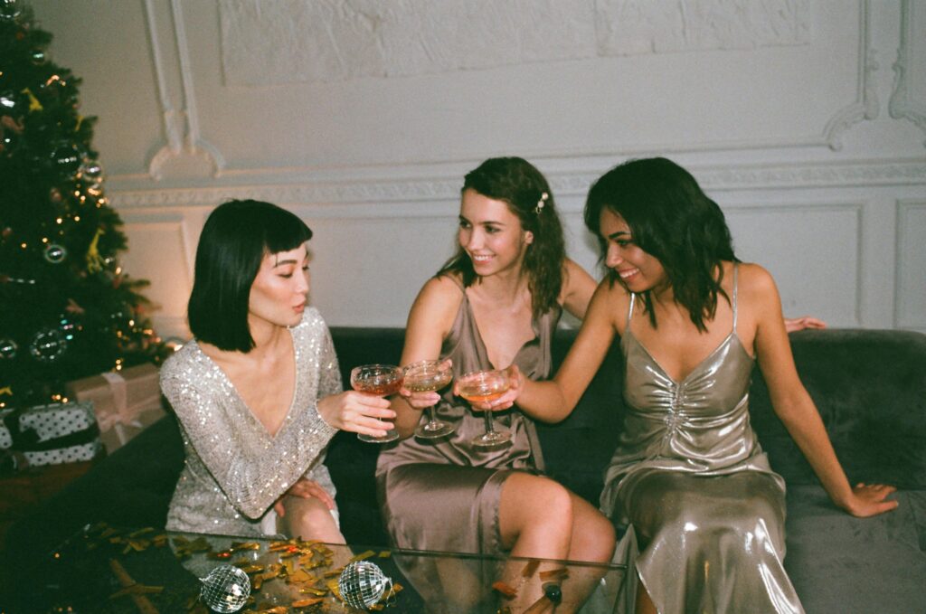 Three friends in glamorous dresses sharing drinks at a festive indoor gathering.